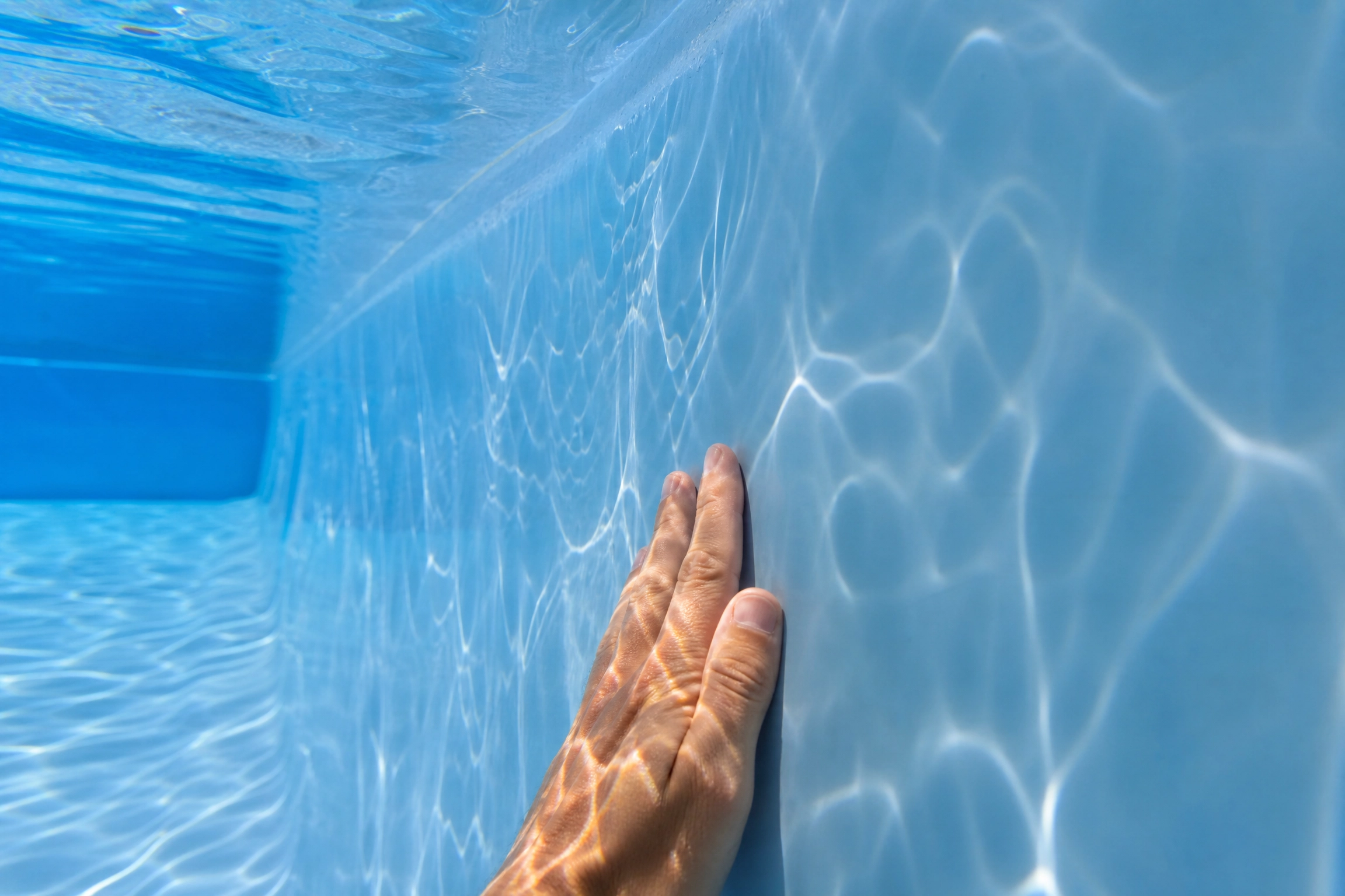 Vue sous-marine d'une main touchant la paroi lisse et intacte d'un bassin, illustrant la durée de vie d'une piscine en fibre de verre bien entretenue